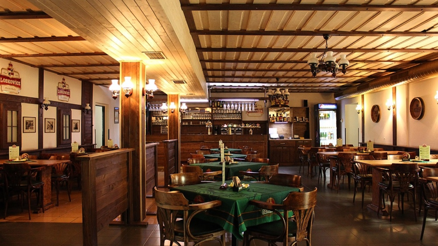 Interiér pivnice — dřevěné stoly, bar a lustry pod trámovým stropem